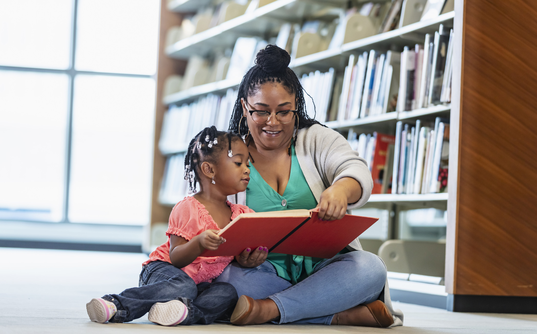 A mixed race mother and daughter sitting on the floor in a library, reading a book. The young child is 3 years old. Her mother is a mature woman in her 40s.